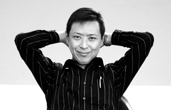 Black and white portrait of Felix Cheong sitting in a chair with his hands folded behind his head and smiling warmly at the camera.