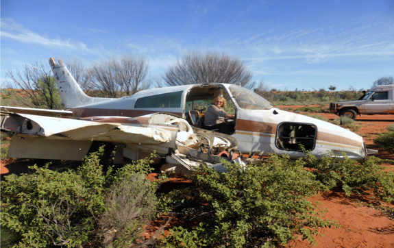 In the middle of a bushy desert, a woman sits inside the cockpit of an aircraft. This aircraft crashed here. No one was killed. 