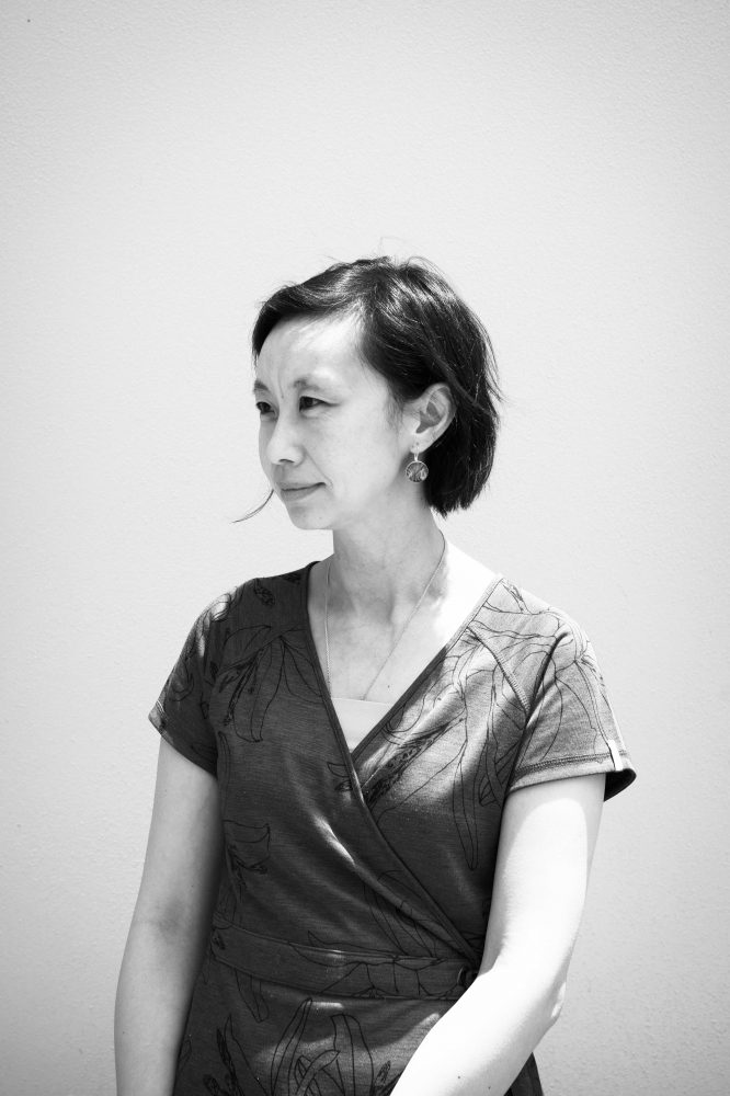 A black and white photo of Emily against a bright orange wall. She is looking off into the distance and is wearing a green dress and green earrings.
