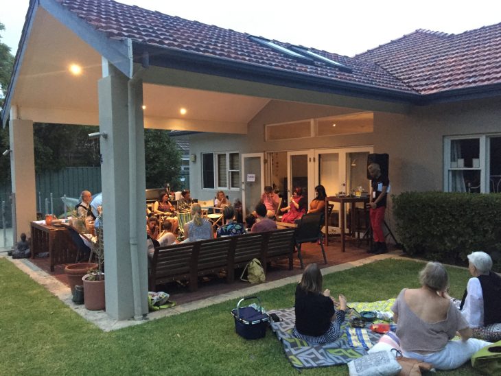 Photo of Nedlands Backstories. Many people sit under a patio and on the lawn in the backyard of a house. Three people sit on chairs in front of the audience. 
