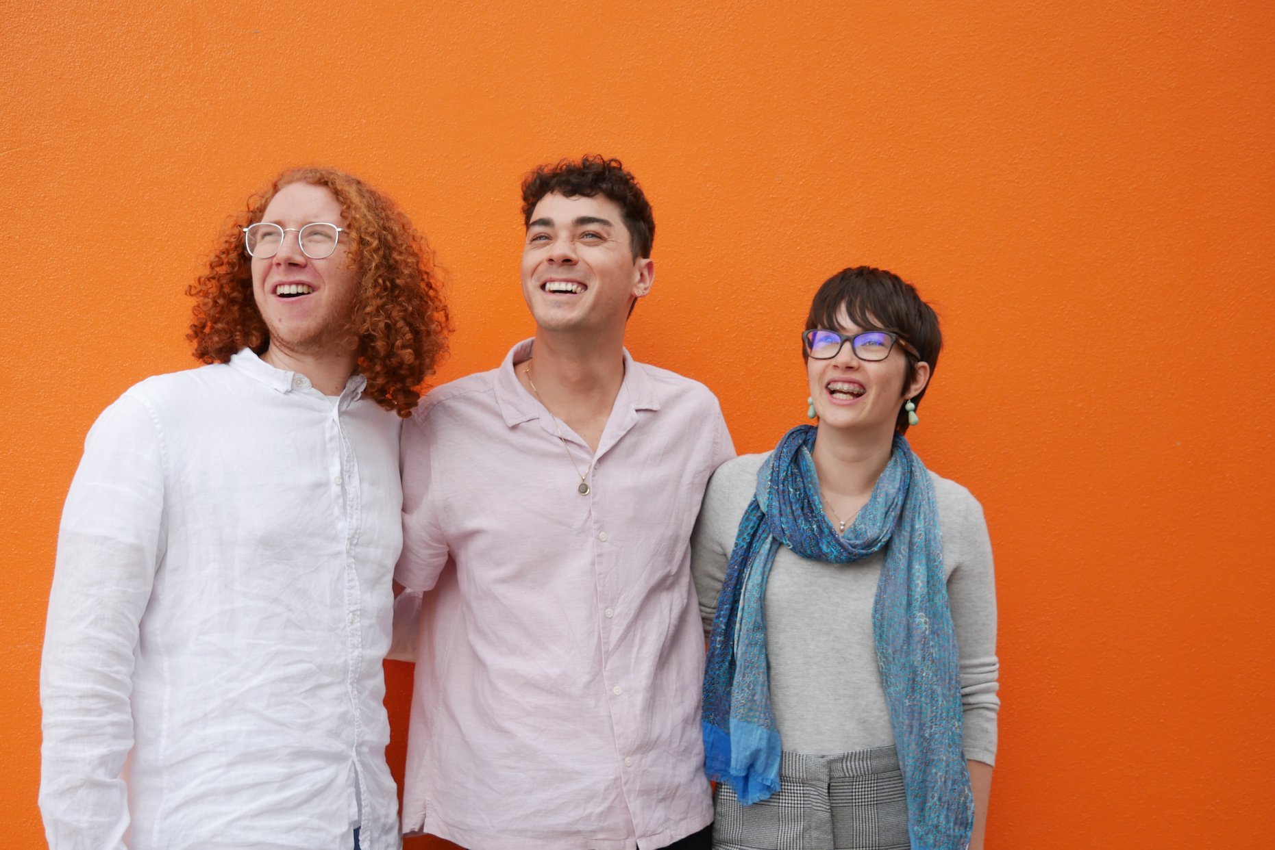 A photograph of three people embracing and smiling. They are all looking past the camera at something to the left. They stand in front of a bright orange wall. The peopple are: Chris Leopardi, Jay Anderson, and Luisa Mitchell