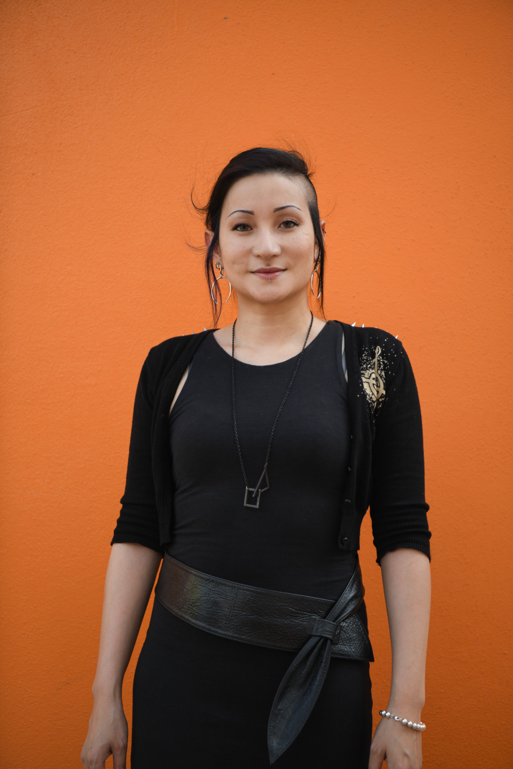 A photograph of Adele Aria standing in front of a bright orange wall. Adele has funky hair and is wearing black.