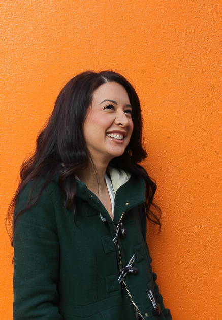 A photograph of Kim Lateef. Kim is standing in front of a bright orange wall and is smiling directly at the camera.