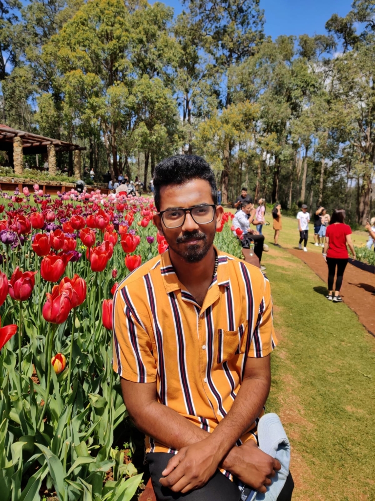 A photograph of Arun sitting beside a bed of tulips and smiling at the camera.