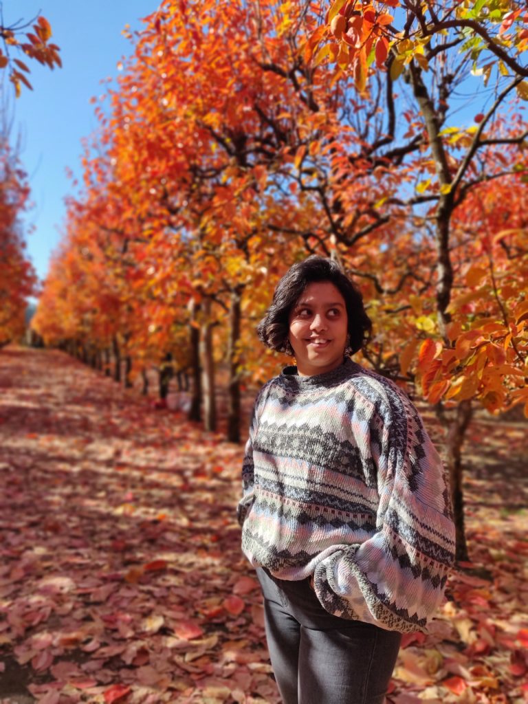 A photograph of Kanchana. They are standing in a beautiful garden of autumn leaves on trees.