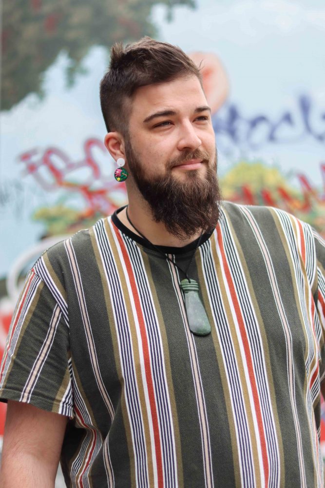 A portrait of Daley Rangi. They wear a stripey top and they are looking off and smiling. The background is a wall with lots of graffiti. 