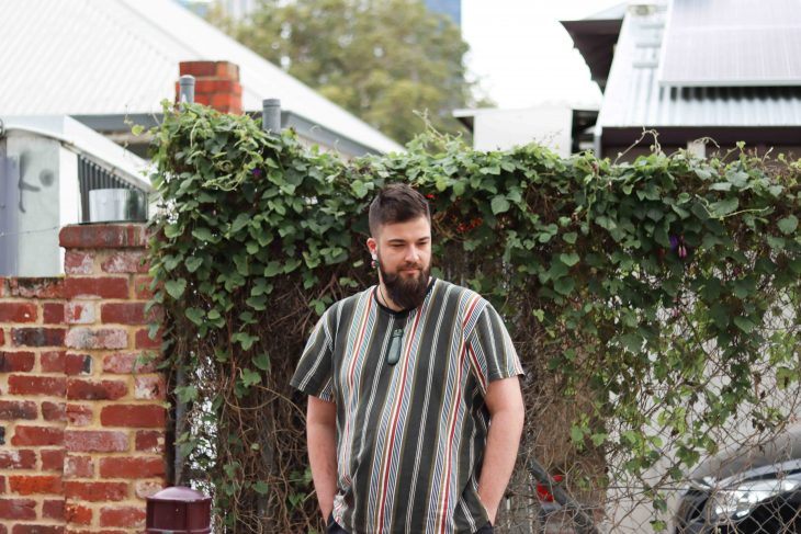 A photograph of Daley Rangi. They are wearing a striped top and they are standing in front of a lucious creeping plant that is covering a wire fence.