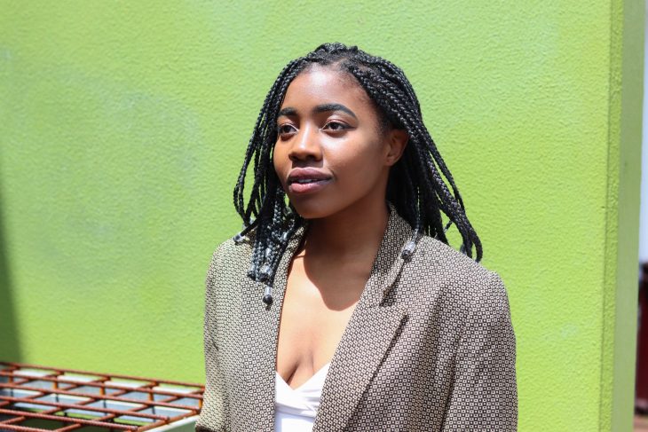 A portrait of Vuma Phiri standing in front of a bright lime green wall. She is looking away from the camera.