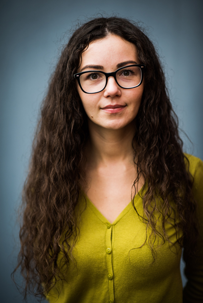 Young woman with long brown curly hair and black framed glasses stands in front of a blue wall and looks away.
