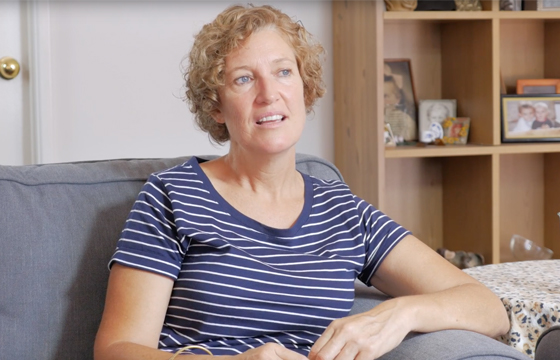 Middle-aged woman with very short curly blonde hair and a stripy navy and white t-shirt sits on a grey couch in a room during the day with a shelf behind her featuring framed family photographs.