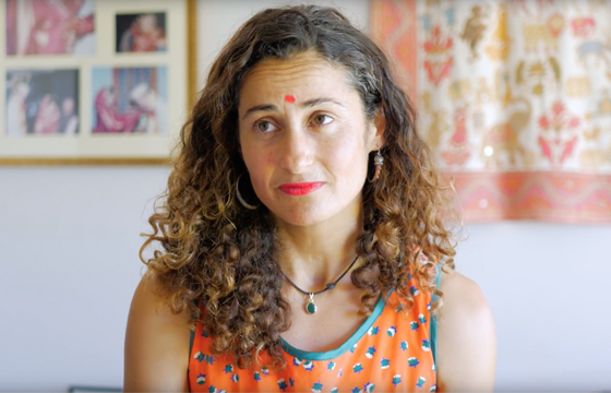 Middle-aged woman with curly brown hair and red bindi on her forehead sits in a lounge with colourful textiles on the walls in the daytime.