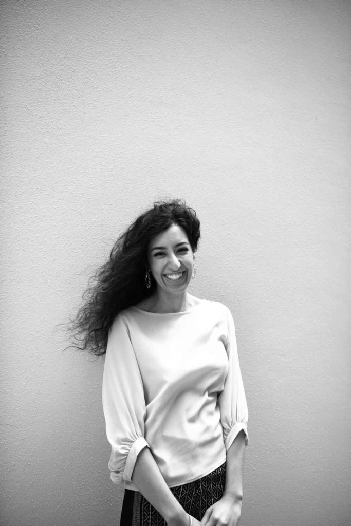 Priya Kahlon is standing before an orange wall. She is laughing and the wind is in her hair.