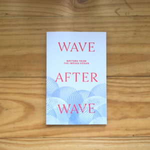 A photograph of a book against a light wood table. The book cover is mostly cream with a blue tattoo-style wave pattern at the bottom. Across the cover in bold red font it says 'WAVE AFTER WAVE'