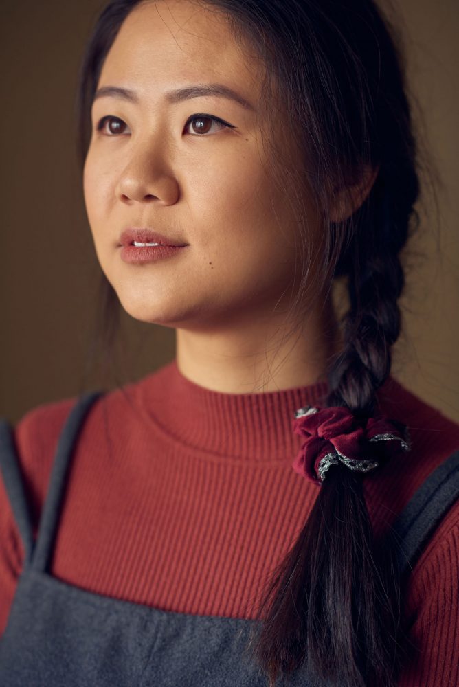 A portrait of Tiffany Ko. Tiffany is wearing a brick-coloured long sleeve top and overalls. She is looking away towards a soft light.