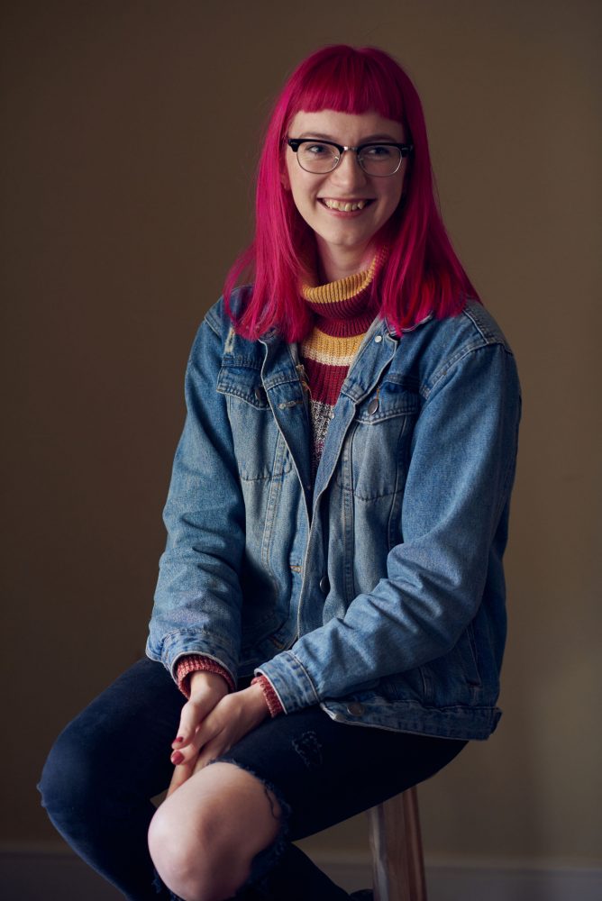 Josephine Newman has pink hair and horn rimmed glasses. She is sitting on a stool and looking at the camera smiling warmly.