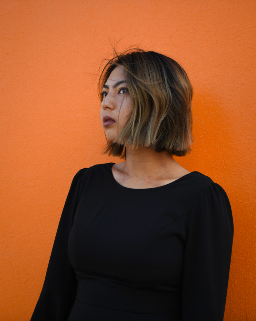 A portrait of Prema Arasu. Prema is photographed against a bright orange wall. They are looking away and the wind is blowing through their hair.