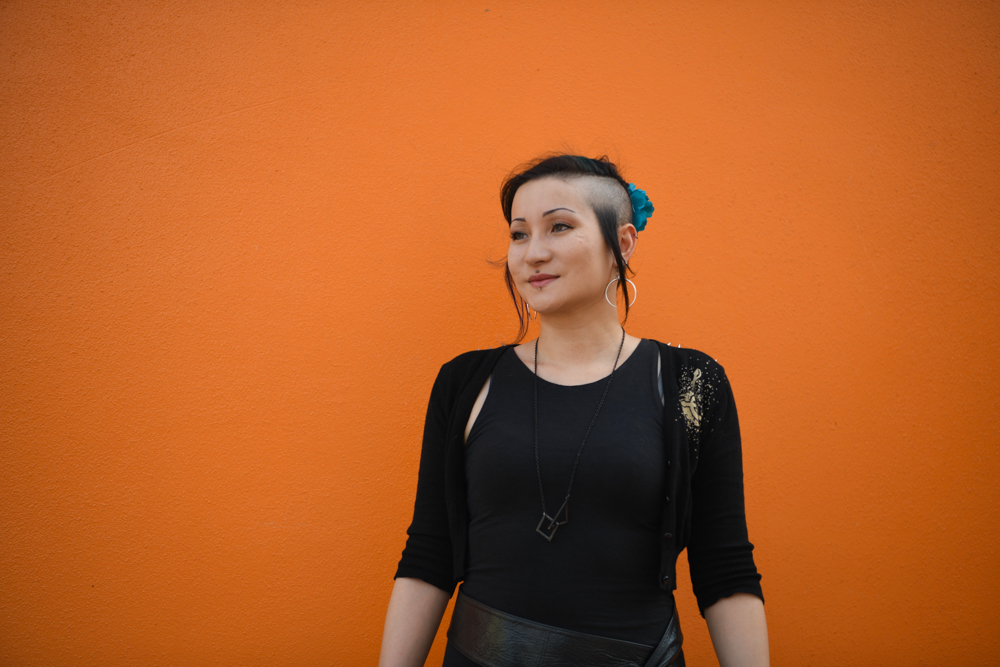 A photograph of Adele Aria standing in front of a bright orange wall. Adele has funky hair and is wearing black. 