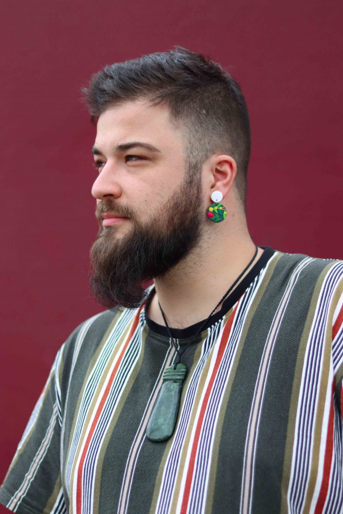 A portrait of Daley Rangi. They are looking away from the camera and they are wearing beautiful circular earrings and a stripy top. They also wear a stunning necklace. The background is a deep maroon colour.