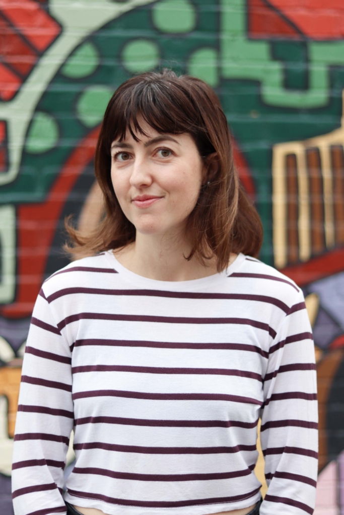 Nadia is standing in front of a colourful background. She is smiling slightly at the camera.