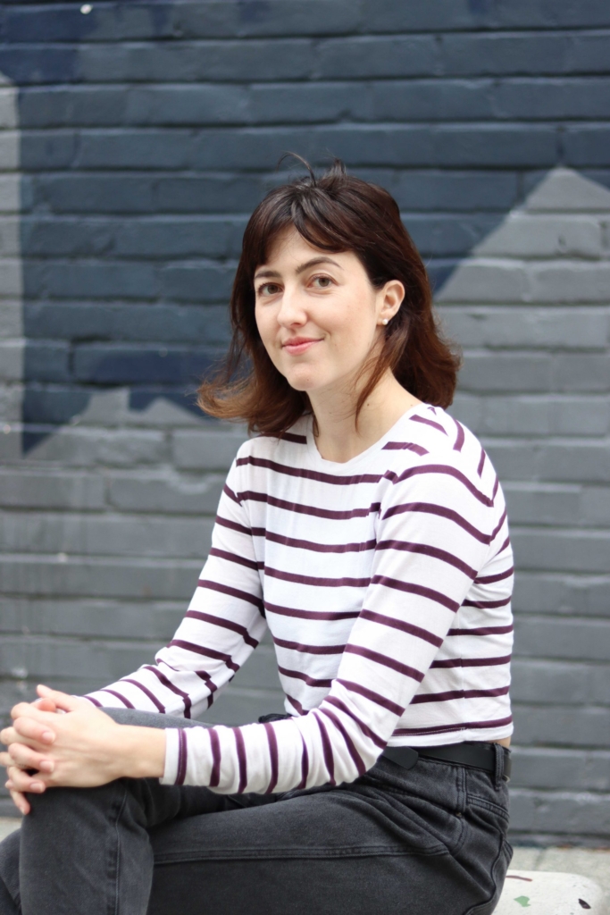 Nadia is sitting in a relaxed manner with her hands on her knees. She is smiling at the camera.