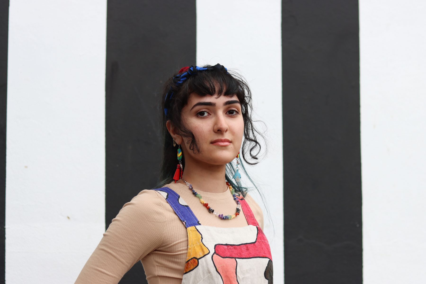 A portrait of Baran standing in front of a vertical striped black and white wall. Baran is wearing very colourful overalls and cool earrings.