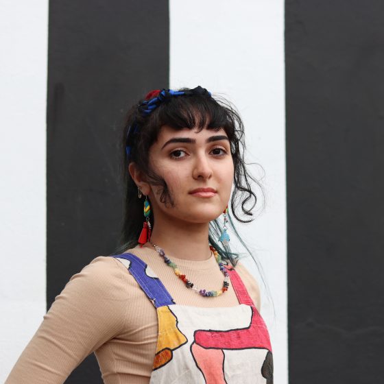 A portrait of Baran standing in front of a vertical striped black and white wall. Baran is wearing very colourful overalls and cool earrings.