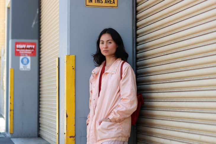Jess Nyanda is standing before a grey wall and a roller door. Jess is wearing a nice light pink jacket and deep red shirt.