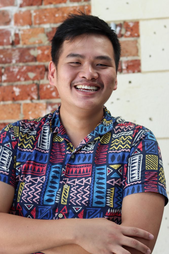 A portrait of William Huang smiling at the camera. He is wearing a funky and bright 80s style button down t shirt