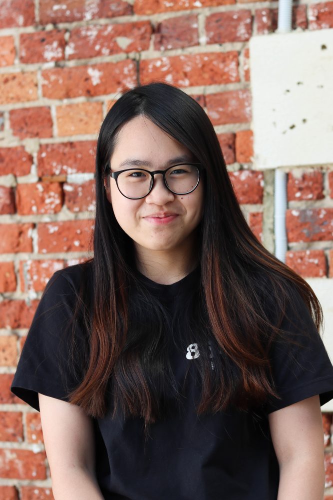 A photo of Maggie Leung. She is wearing glasses and smiling at the camera. Behind her is a red brick wall.