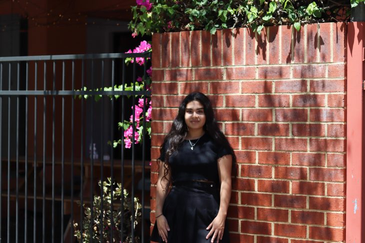 A portrait of Camila. She is standing in front of a red brick wall and smiling. Camila is wearing all black. Behind her on the left is pink flowers.
