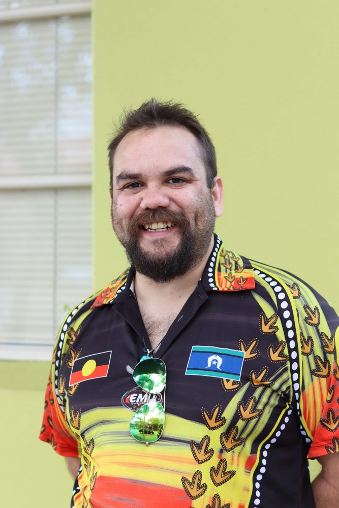 A very smiley portrait of Daniel Hansen. He is standing in front of a lime green wall with his hands resting by his side. He is wearing a shirt that is black, red, yellow, orange and green full of Aboriginal art. The shirt also has an Aboriginal flag and a Torres Strait Islander flag on it.