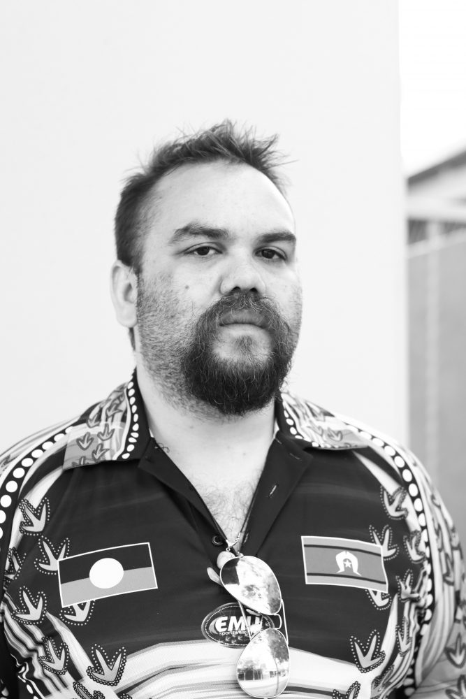 A black and white photograph of Daniel Hansen. He is standing looking calmly at the camera. He is wearing a very fun shirt that has Aboriginal art all over it. There is also an Aboriginal flag and a Torres Strait Islander flag on the shirt.