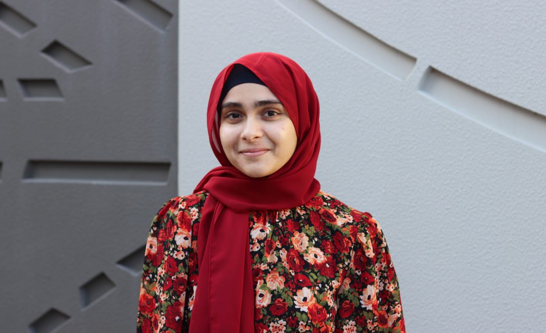 A portrait of Zahina. She is wearing a beautiful red head scarf and a matching floral dress full of red, yellow and orange flowers. She is smiling at the camera.