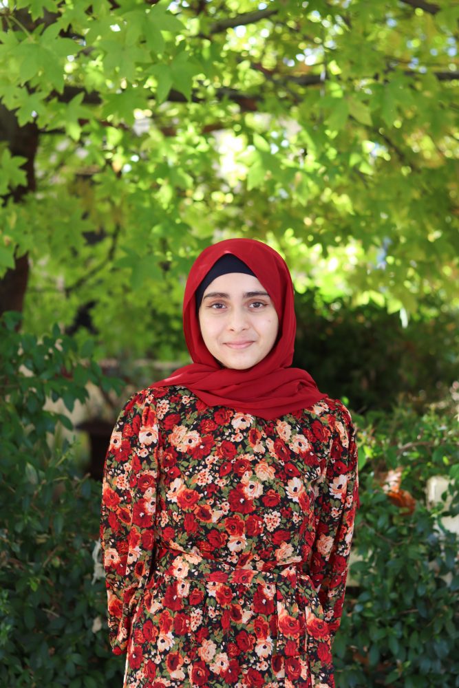 A portrait of Zahina standing in the streets of Northbridge. She is wearing a beautiful red head scarf and a matching floral dress full of red, yellow and orange flowers. She is smiling at the camera.