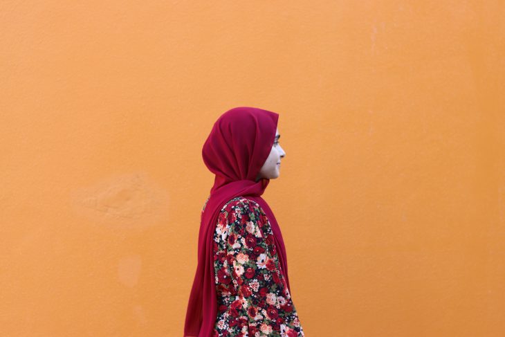 A portrait of Zahina. She is standing facing the right of the camera, looking off into the distance. Behind her is a brilliant orange wall.