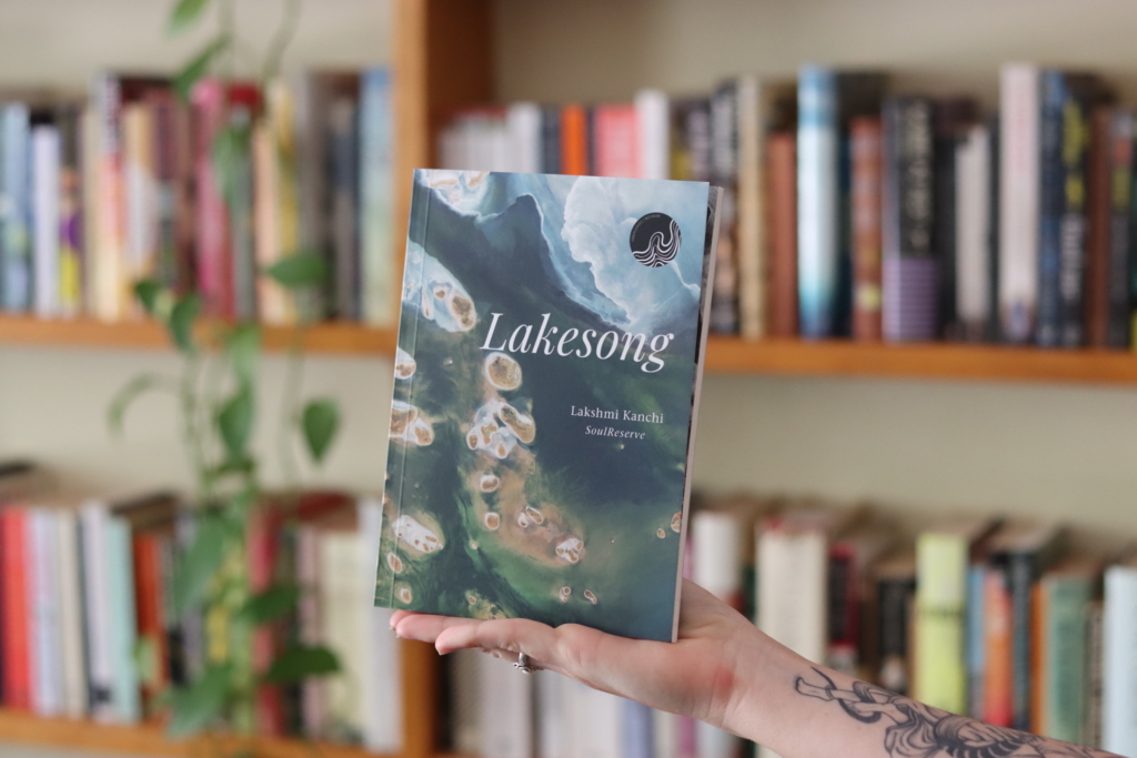 A photograph of a book held the the palm of a person's hand. In the background is a beautiful book shelf. The book cover is an aerial shot of a lake with the title Lakesong in white at the top.