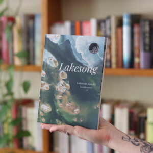 A photograph of a book held the the palm of a person's hand. In the background is a beautiful book shelf. The book cover is an aerial shot of a lake with the title Lakesong in white at the top.