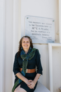 A photo of Jo sitting on a wall next to a sign. On the sign it says 'this property was donated to the Salvation Army by Mrs E A Pearse. For the purposes of Maternity Hospital.' This hospital is where baby Jo was taken from her mother. 