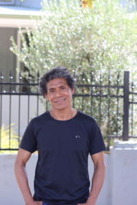 A portrait of Jose da Costa. He is standing outside in the shade of a Mulberry tree. Behind him is a fence with an olive tree. Jose is wearing a black t-shirt and is smiling at the camera with his hands in his pocket. 