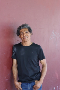 A portrait of Jose da Costa. He is standing in front of a solid red-brown wall outside. He is wearing a black t-shirt and is smiling at the camera. He has his hands in his jeans pockets. 