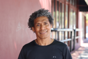 A portrait of Jose da Costa. He is standing in front of a solid red-brown wall outside. He is wearing a black t-shirt and is smiling at the camera.